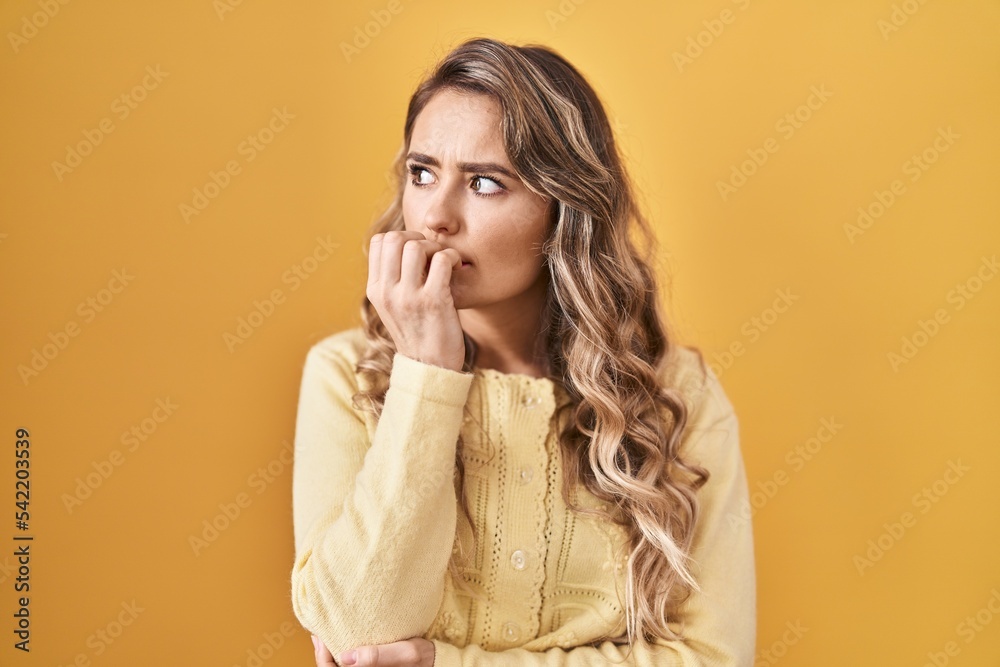 Young caucasian woman standing over yellow background looking stressed and nervous with hands on mouth biting nails. anxiety problem. Young caucasian woman standing over yellow background looking stressed and nervous with hands on mouth biting nails. anxiety problem.