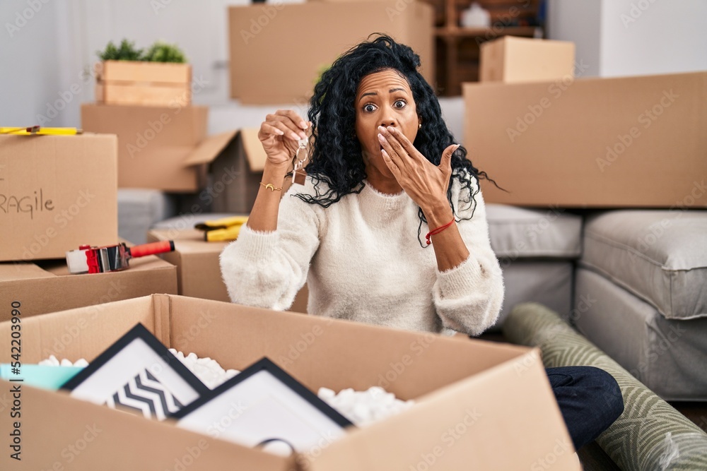 Middle age hispanic woman holding keys of new home covering mouth with hand, shocked and afraid for mistake. surprised expression Middle age hispanic woman holding keys of new home covering mouth with hand, shocked and afraid for mistake. surprised expression