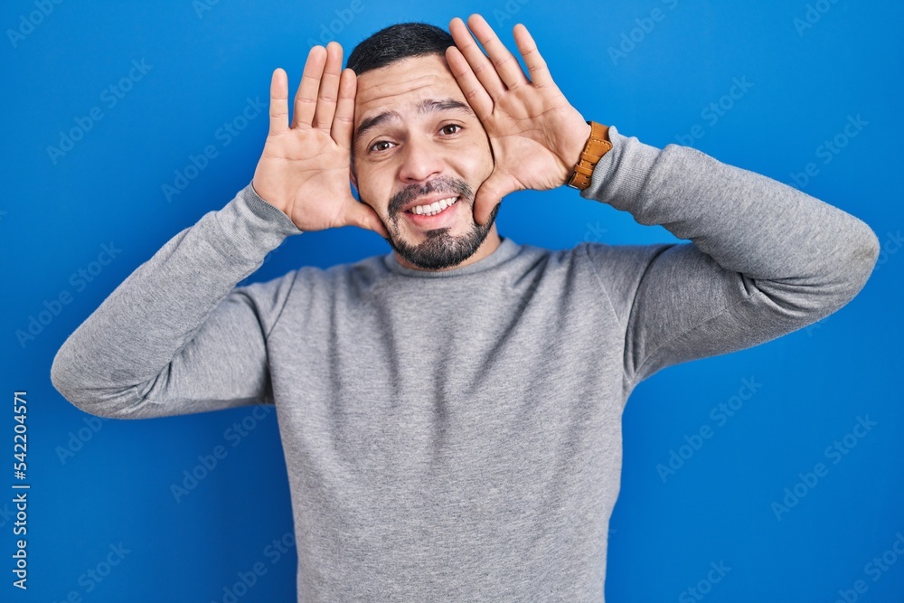 Hispanic man standing over blue background smiling cheerful playing peek a boo with hands showing face. surprised and exited Hispanic man standing over blue background smiling cheerful playing peek a boo with hands showing face. surprised and exited