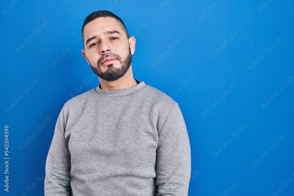 Hispanic man standing over blue background relaxed with serious expression on face. simple and natural looking at the camera. Hispanic man standing over blue background relaxed with serious expression on face. simple and natural looking at the camera.