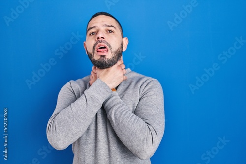 Hispanic man standing over blue background shouting suffocate because painful strangle. health problem. asphyxiate and suicide concept.