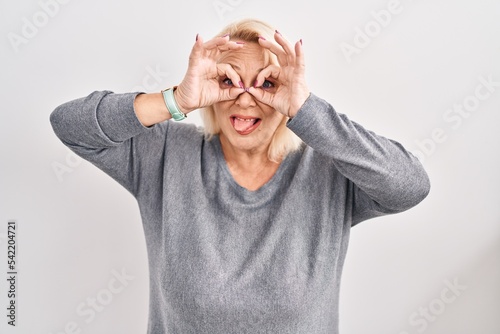 Middle age caucasian woman standing over white background doing ok gesture like binoculars sticking tongue out, eyes looking through fingers. crazy expression.