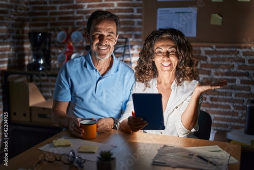 Middle age hispanic couple using touchpad sitting on the table at night smiling cheerful presenting and pointing with palm of hand looking at the camera.