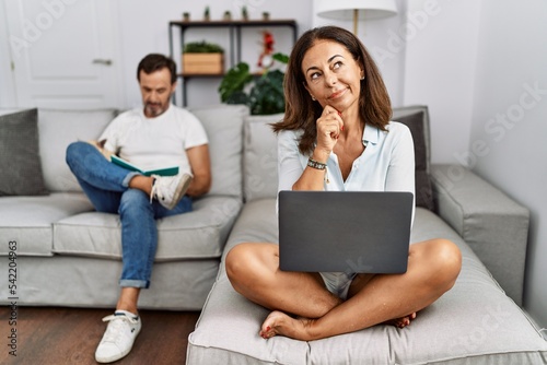 Hispanic middle age couple at home, woman using laptop looking confident at the camera smiling with crossed arms and hand raised on chin. thinking positive.