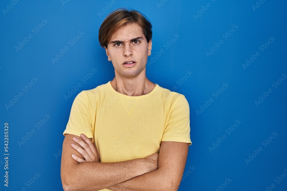 Young man standing over blue background skeptic and nervous, disapproving expression on face with crossed arms. negative person. Young man standing over blue background skeptic and nervous, disapproving expression on face with crossed arms. negative person.