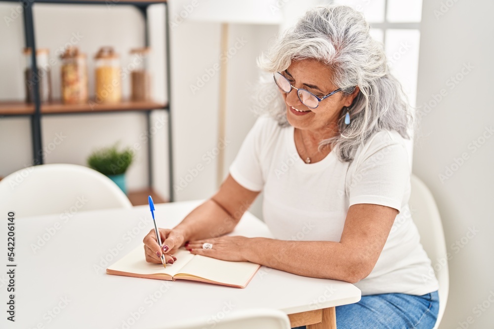Middle age woman writing on notebook sitting on table at home