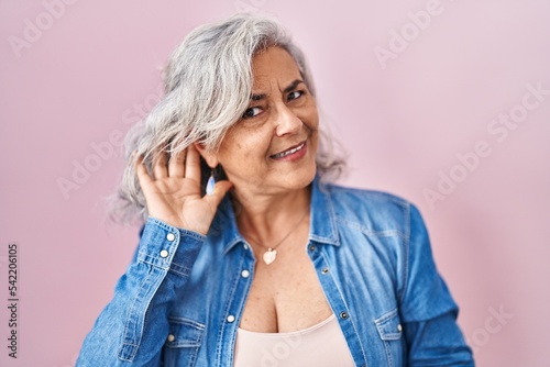 Middle age woman with grey hair standing over pink background smiling with hand over ear listening an hearing to rumor or gossip. deafness concept. photo