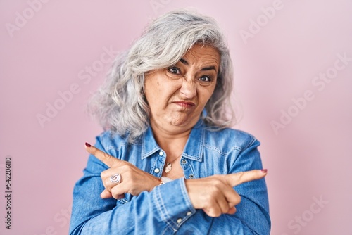 Middle age woman with grey hair standing over pink background pointing to both sides with fingers, different direction disagree