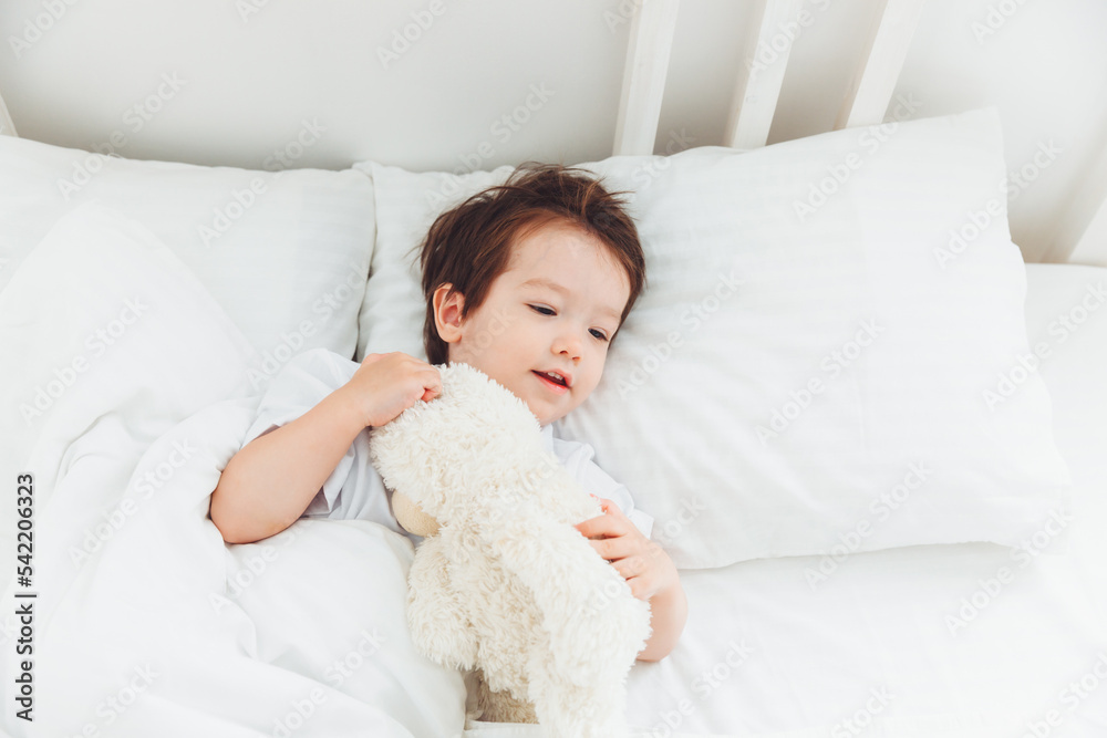 A little Asian boy in pajamas lying on the bed with a teddy bear, The child wakes up happy, The child wakes up early in the morning.natural bed linen for children A little Asian boy in pajamas lying on the bed with a teddy bear, The child wakes up happy, The child wakes up early in the morning.natural bed linen for children