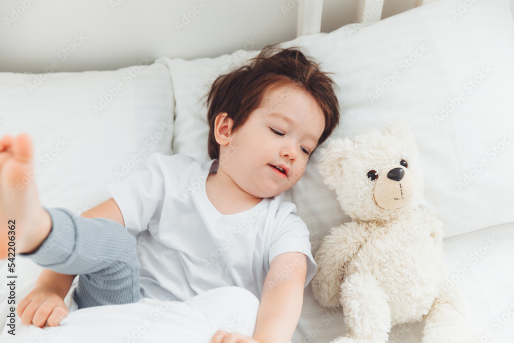 A little Asian boy in pajamas lying on the bed with a teddy bear, The child wakes up happy, The child wakes up early in the morning.natural bed linen for children A little Asian boy in pajamas lying on the bed with a teddy bear, The child wakes up happy, The child wakes up early in the morning.natural bed linen for children