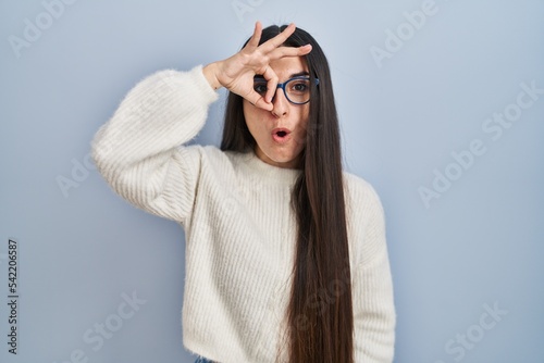 Young hispanic woman wearing casual sweater over blue background doing ok gesture shocked with surprised face, eye looking through fingers. unbelieving expression.
