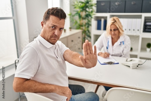 Hispanic man at the doctor with open hand doing stop sign with serious and confident expression, defense gesture
