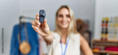 Young blonde woman smiling confident holding clothes security alarm at clothing store