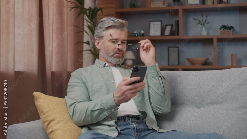 Cheerful middle aged man with grey hair sitting on the sofa using phone. Smiling wearing glasses in the modern apartment. Texting message scrolling. Portrait Cheerful middle aged man with grey hair sitting on the sofa using phone. Smiling wearing glasses in the modern apartment. Texting message scrolling. Portrait