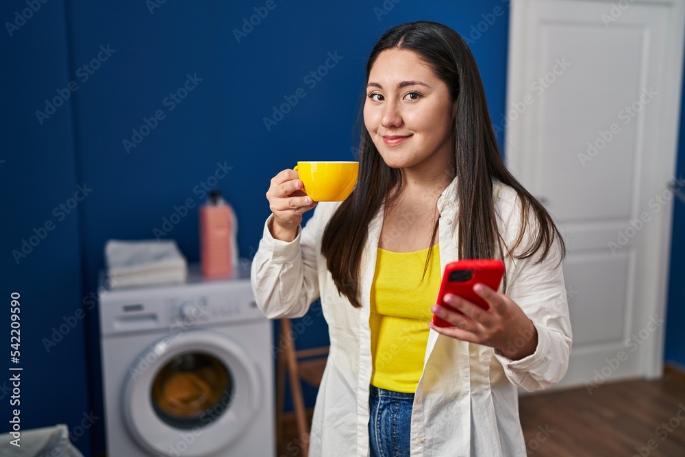 Young hispanic woman using smartphone and drinking coffee waiting for washing machine at laundry room Young hispanic woman using smartphone and drinking coffee waiting for washing machine at laundry room