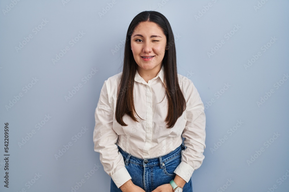 Young latin woman standing over blue background winking looking at the camera with sexy expression, cheerful and happy face. Young latin woman standing over blue background winking looking at the camera with sexy expression, cheerful and happy face.