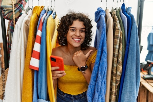 Young hispanic woman searching clothes on clothing rack using smartphone pointing to the back behind with hand and thumbs up, smiling confident