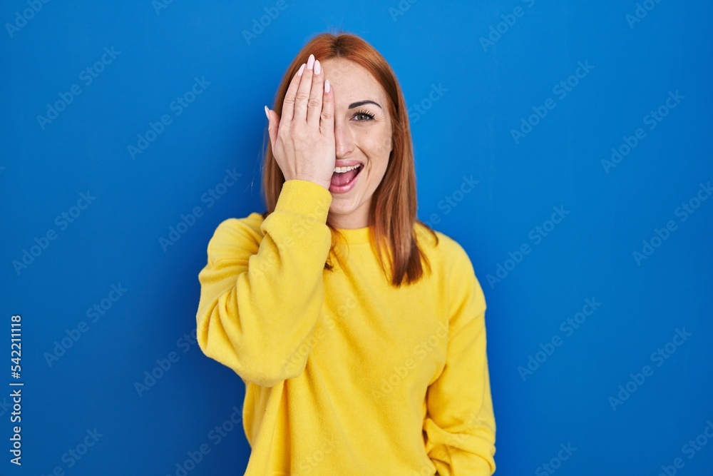 Young woman standing over blue background covering one eye with hand, confident smile on face and surprise emotion. Young woman standing over blue background covering one eye with hand, confident smile on face and surprise emotion.