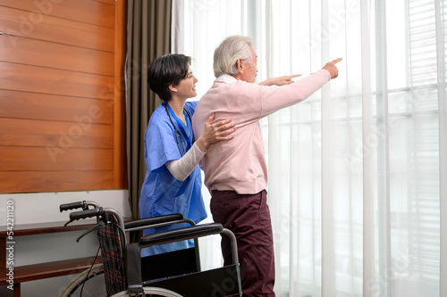 Asian nurse taking care of an elderly man getting up from wheelchair at senior healthcare center.