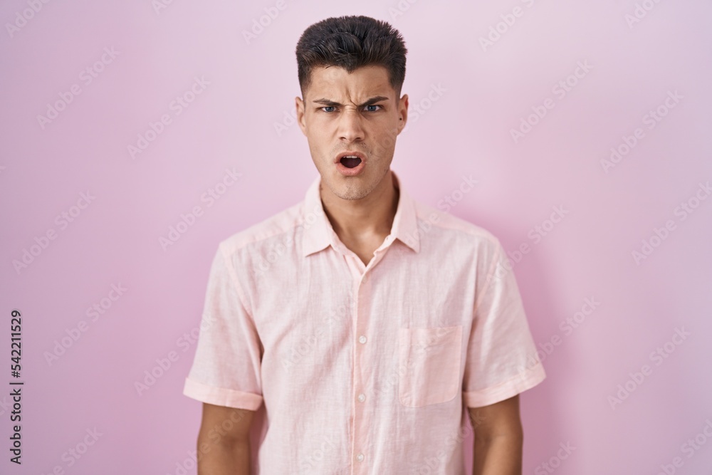 Young hispanic man standing over pink background in shock face, looking skeptical and sarcastic, surprised with open mouth Young hispanic man standing over pink background in shock face, looking skeptical and sarcastic, surprised with open mouth