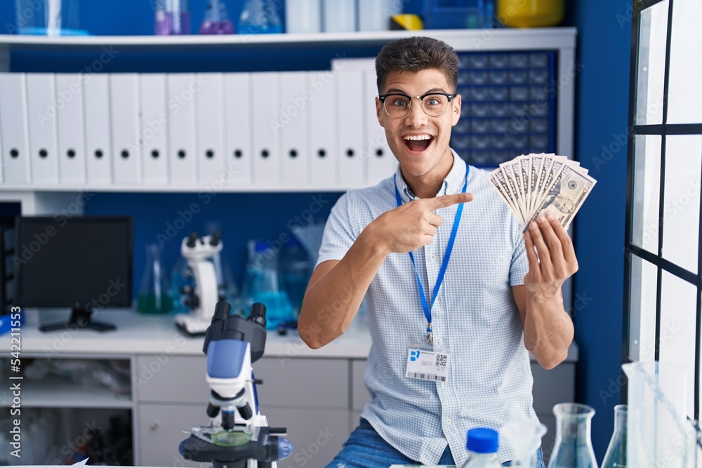 Young hispanic man working at scientist laboratory holding dollars smiling happy pointing with hand and finger Young hispanic man working at scientist laboratory holding dollars smiling happy pointing with hand and finger