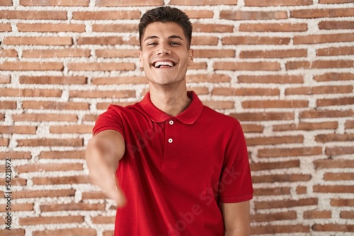 Young hispanic man standing over bricks wall smiling friendly offering handshake as greeting and welcoming. successful business.