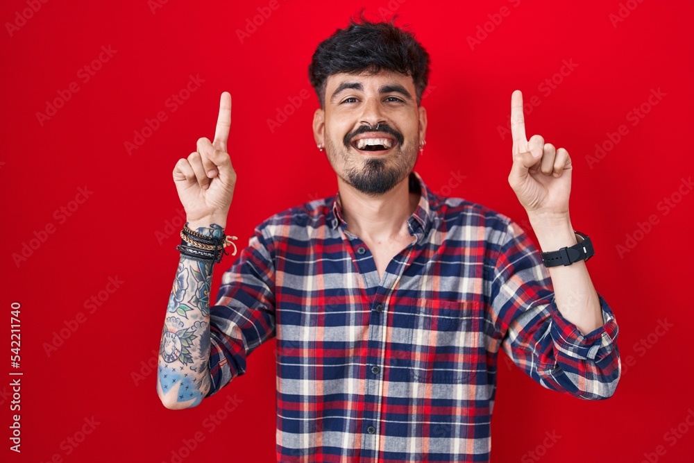 Young hispanic man with beard standing over red background smiling amazed and surprised and pointing up with fingers and raised arms. Young hispanic man with beard standing over red background smiling amazed and surprised and pointing up with fingers and raised arms.
