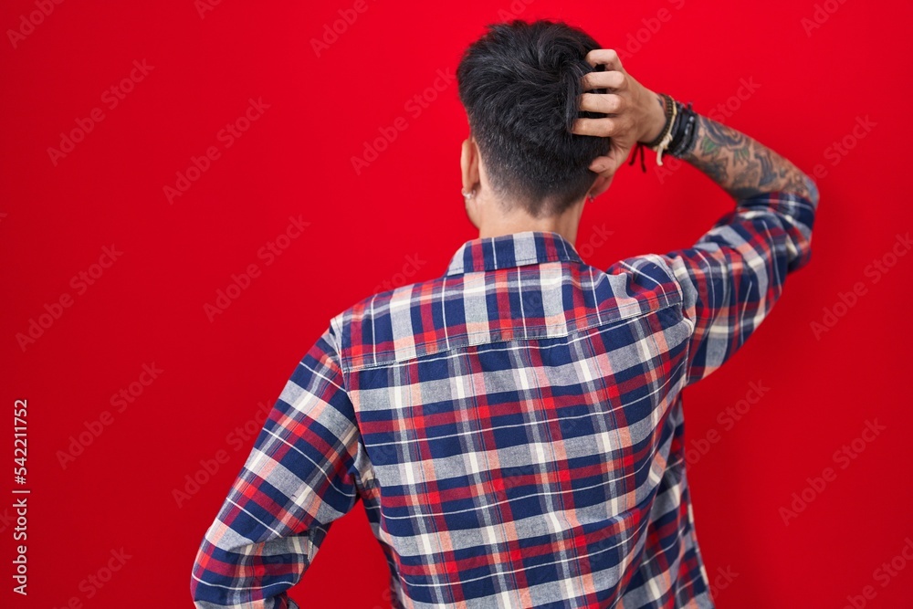 Young hispanic man with beard standing over red background backwards thinking about doubt with hand on head Young hispanic man with beard standing over red background backwards thinking about doubt with hand on head