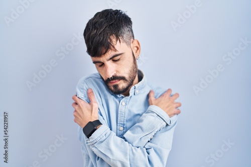 Young hispanic man with beard standing over blue background hugging oneself happy and positive, smiling confident. self love and self care