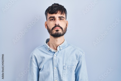 Young hispanic man with beard standing over blue background relaxed with serious expression on face. simple and natural looking at the camera.