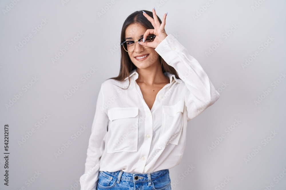 Young brunette woman wearing glasses doing ok gesture with hand smiling, eye looking through fingers with happy face.