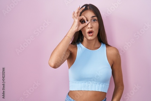 Young brunette woman standing over pink background doing ok gesture shocked with surprised face, eye looking through fingers. unbelieving expression. © Krakenimages.com