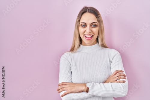 Young blonde woman wearing white sweater over pink background happy face smiling with crossed arms looking at the camera. positive person.