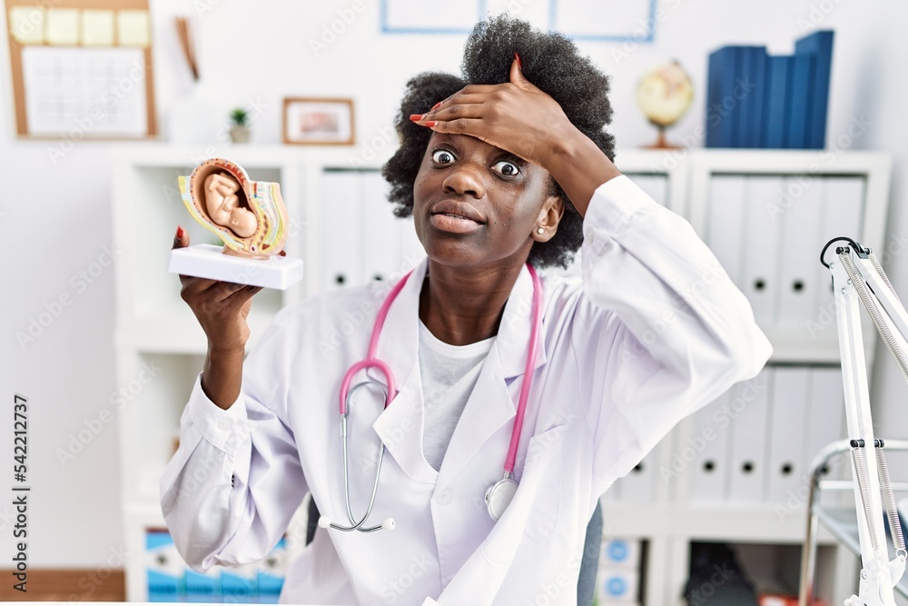 Obraz premium African doctor woman holding anatomical model of female uterus with fetus stressed and frustrated with hand on head, surprised and angry face