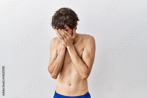 Young hispanic man standing shirtless over isolated, background with sad expression covering face with hands while crying. depression concept.