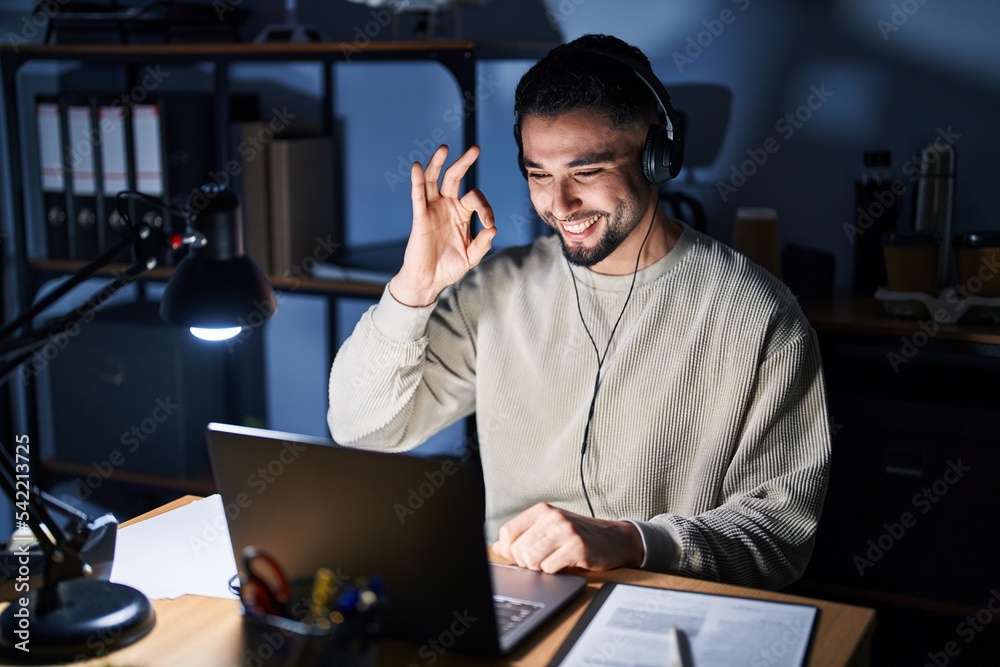 Young handsome man working using computer laptop at night smiling positive doing ok sign with hand and fingers. successful expression.