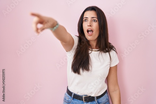 Young brunette woman standing over pink background pointing with finger surprised ahead, open mouth amazed expression, something on the front