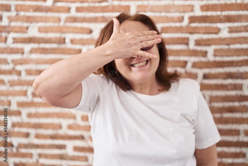 Senior woman with glasses standing over bricks wall smiling and laughing with hand on face covering eyes for surprise. blind concept. Senior woman with glasses standing over bricks wall smiling and laughing with hand on face covering eyes for surprise. blind concept.