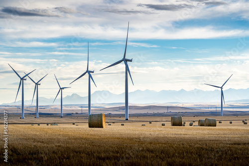 Windmills standing tall on agriculture fields producing alternative energy with round hay bales and Canadian Rocky Mountains at background near Pincher Creek Alberta Canada.