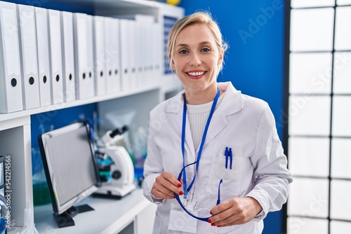 Young blonde woman scientist smiling confident holding glasses at laboratory