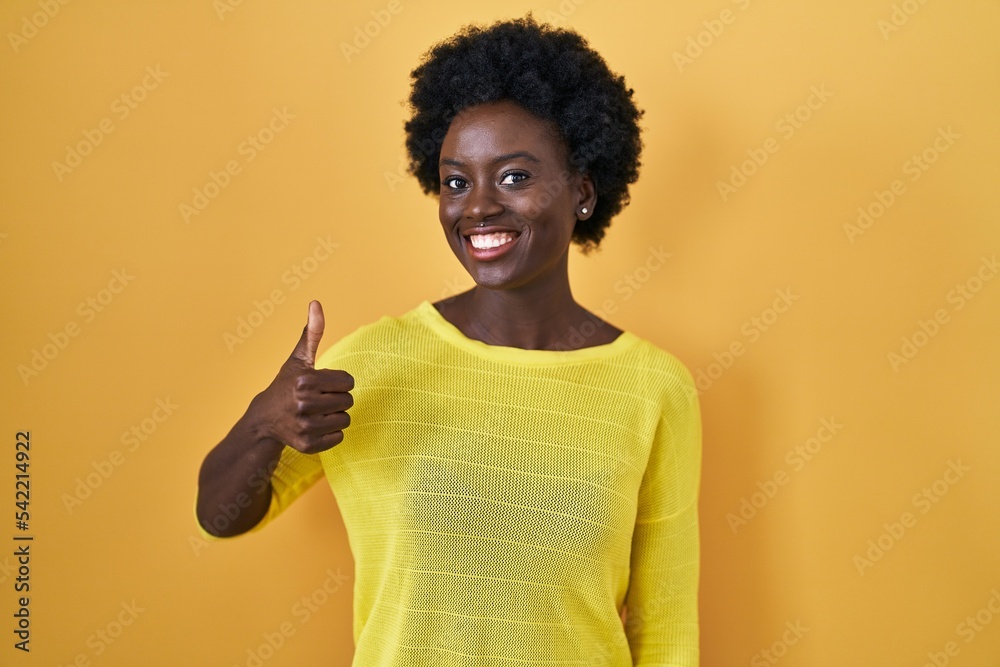 African young woman standing over yellow studio doing happy thumbs up gesture with hand. approving expression looking at the camera showing success. African young woman standing over yellow studio doing happy thumbs up gesture with hand. approving expression looking at the camera showing success.
