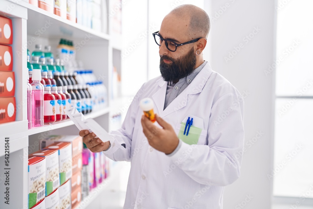 Young bald man pharmacist holding pills bottle reading prescription at pharmacy