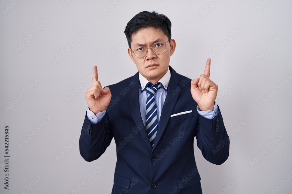 Young asian man wearing business suit and tie pointing up looking sad and upset, indicating direction with fingers, unhappy and depressed. Young asian man wearing business suit and tie pointing up looking sad and upset, indicating direction with fingers, unhappy and depressed.