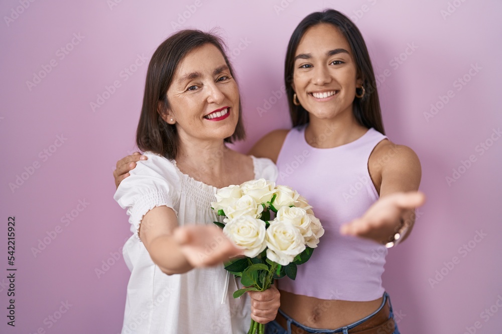 Hispanic mother and daughter holding bouquet of white flowers smiling cheerful offering palm hand giving assistance and acceptance.