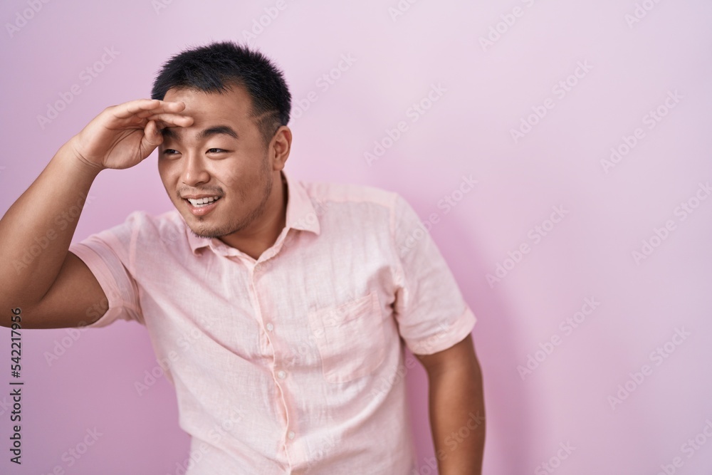 Chinese young man standing over pink background very happy and smiling looking far away with hand over head. searching concept. Chinese young man standing over pink background very happy and smiling looking far away with hand over head. searching concept.