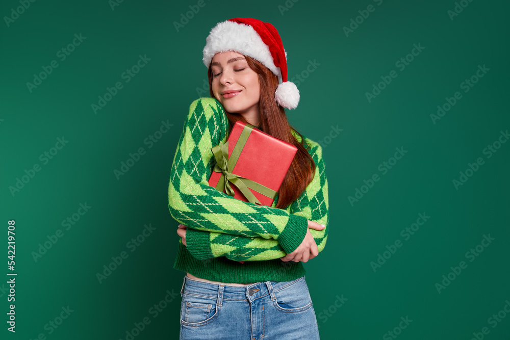 Attractive young woman in Christmas hat carrying gift box and smiling against green background Attractive young woman in Christmas hat carrying gift box and smiling against green background