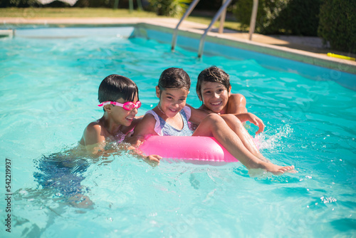 Portrait of smiling children having fun in pool. Two little Caucasian boys standing in water and beautiful girl sitting on inflatable ring looking straight. Kids active rest, summer vacation concept