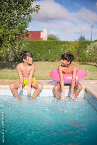 Portrait of two Caucasian boys sitting together on edge of pool. Smiling cute kids resting with their legs in water splashing it looking at each other with joy. Leisure activity and friendship concept