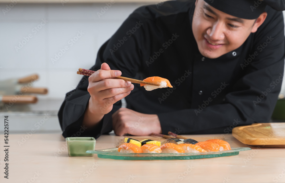 Selective focus on delicious fresh salmon sushi in chopsticks in hand of young handsome Asian chef with black uniform and hat, preparing Japanese food, at restaurant kitchen Selective focus on delicious fresh salmon sushi in chopsticks in hand of young handsome Asian chef with black uniform and hat, preparing Japanese food, at restaurant kitchen