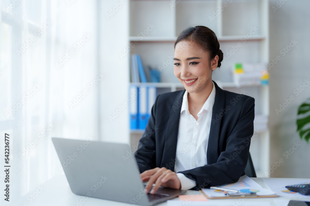 Happy Asian businesswoman sitting in the office with her laptop computer working diligently and smiling brightly with loads of papers on her desk. Happy Asian businesswoman sitting in the office with her laptop computer working diligently and smiling brightly with loads of papers on her desk.